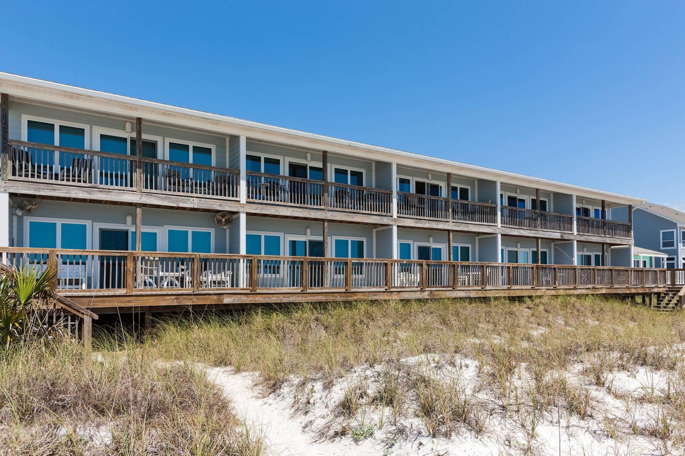 Un hotel frente al mar con varios pisos, balcones, una terraza de madera y arena a su alrededor.