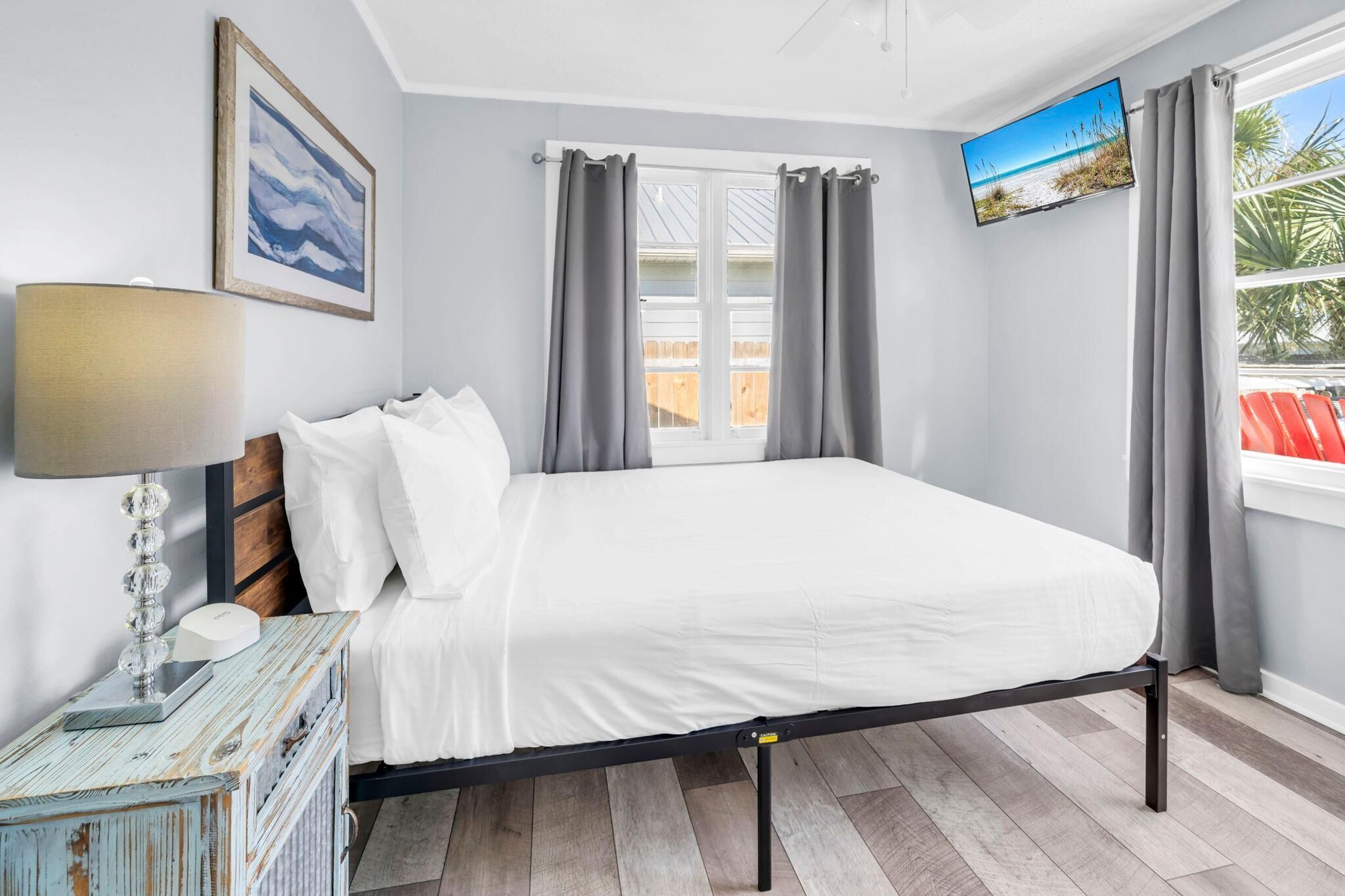 Une chambre à coucher avec un lit, une télévision, une lampe, un tableau et une fenêtre avec des rideaux.