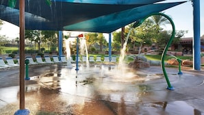 Ein Wasserspiel mit mehreren Düsen, eine überdachte Sitzgelegenheit mit Stühlen und ein gepflasterter Boden.