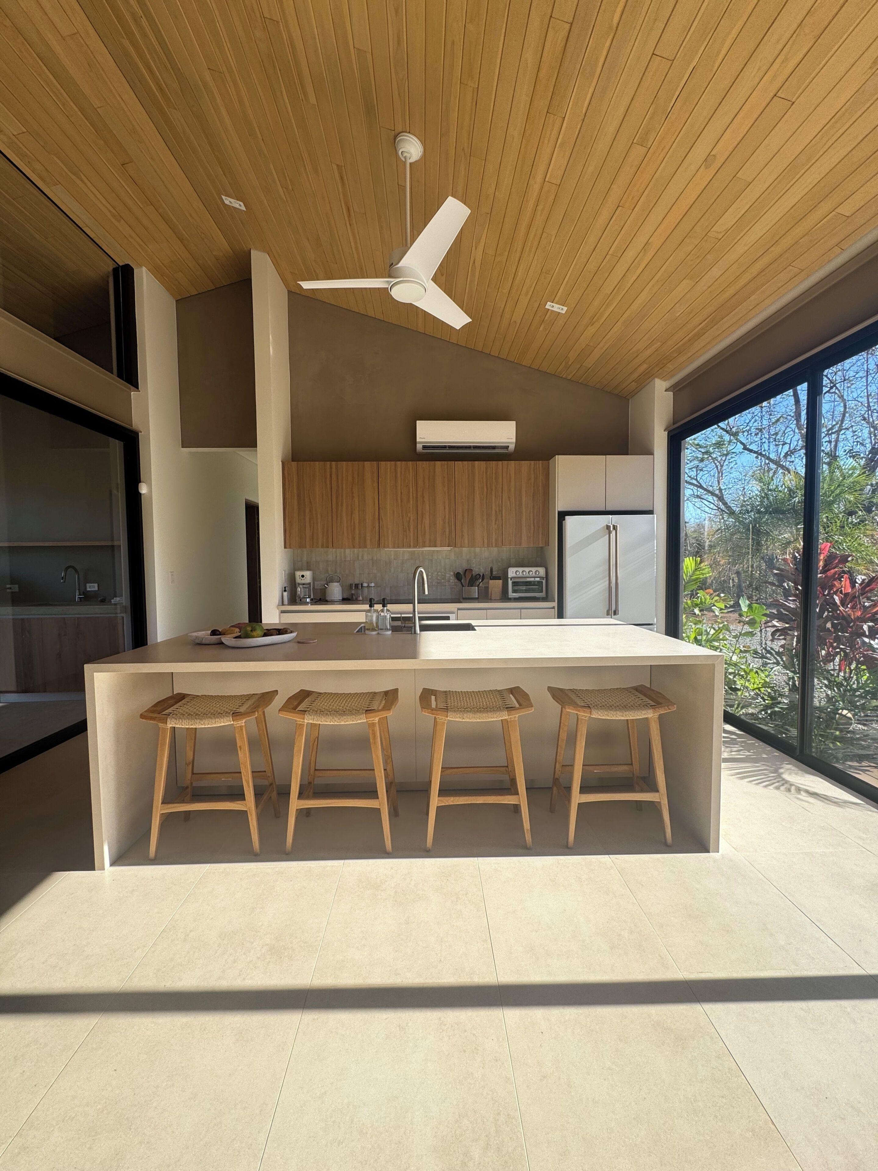 A modern kitchen with a central island, bar stools, a ceiling fan, and a large window providing a view of the outdoors.