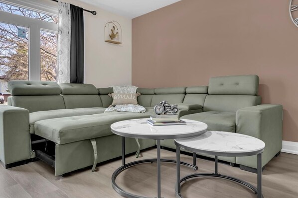 Un canapé vert, deux tables rondes, une horloge et un élément de décoration fixé au mur, dans une pièce dotée d’une fenêtre.