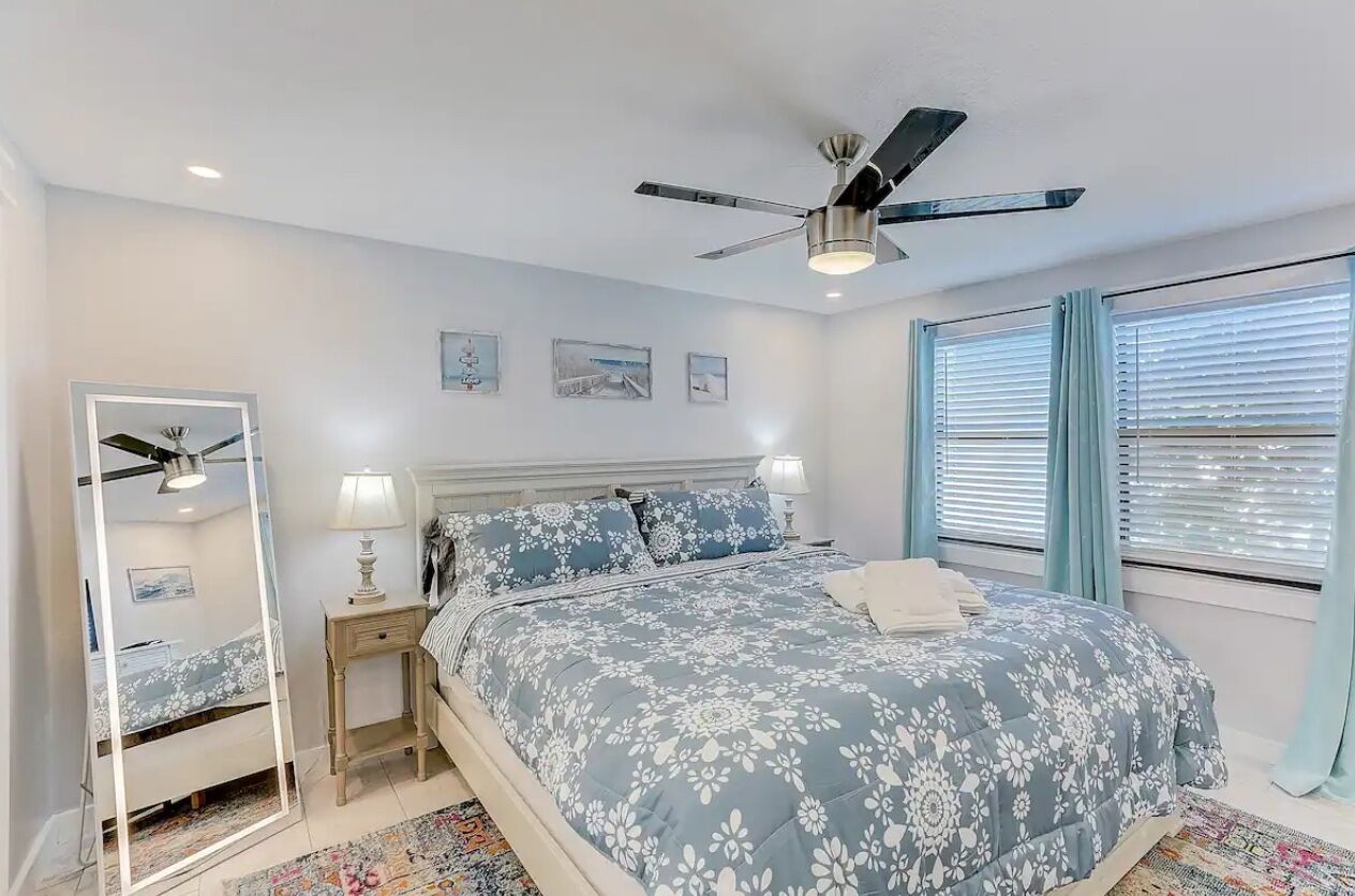 A bedroom with a bed, a ceiling fan, a nightstand, a mirror, and a window with curtains.
