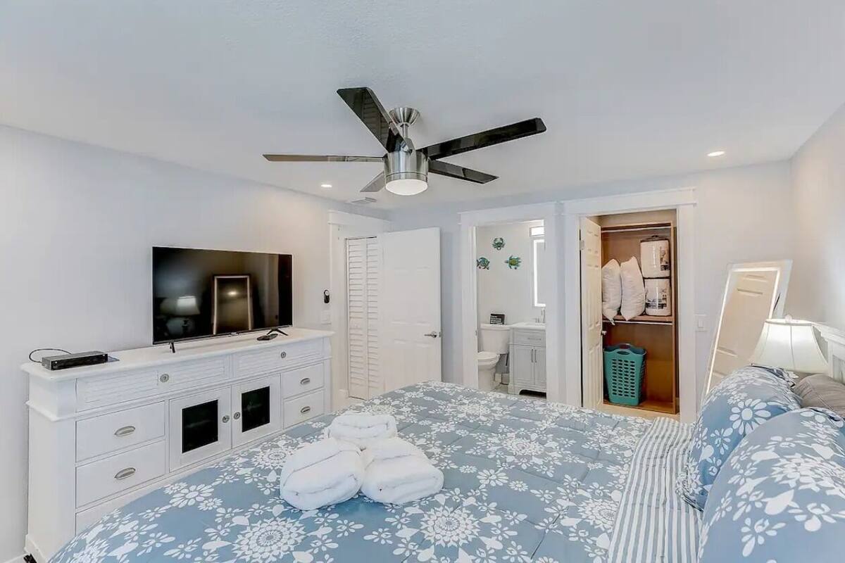 A bedroom with a bed, a TV, a wardrobe, a ceiling fan, and a bathroom visible through an open door.