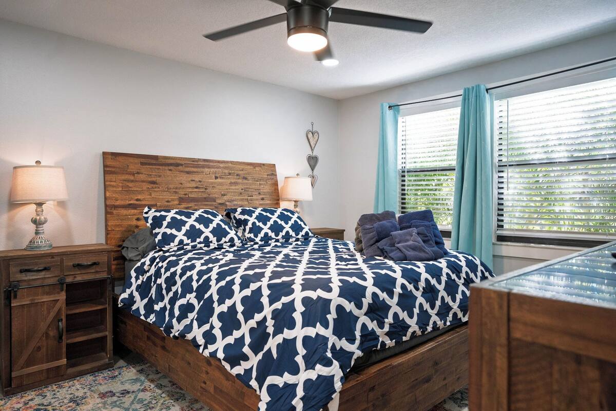 A bedroom with a wooden headboard, a bed with a patterned bedspread, a nightstand with a lamp, a dresser, and a window with blinds.