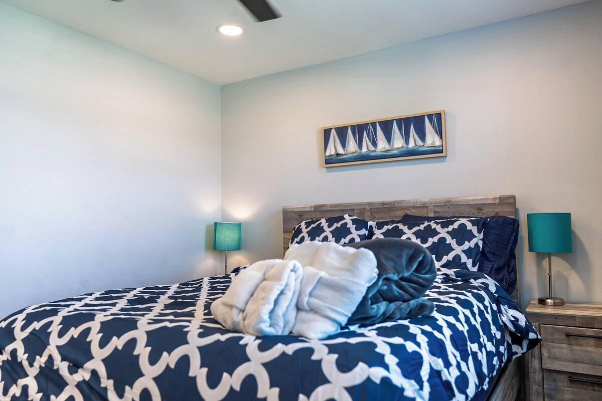 A bed with a patterned bedspread, a headboard, a wall-mounted painting of sailboats, and two bedside lamps.