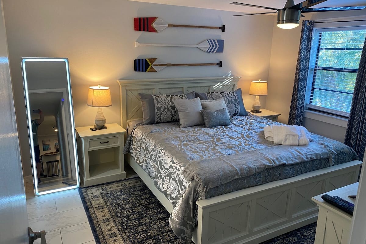 A bedroom with a bed, nightstands, a mirror, a ceiling fan, and a window with curtains.