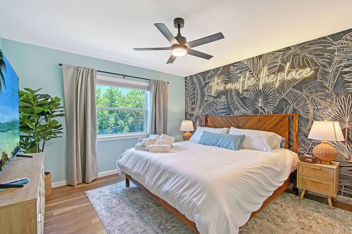 A bedroom with a bed, a ceiling fan, a nightstand with a lamp, a window with curtains, and a wall with a leaf pattern.
