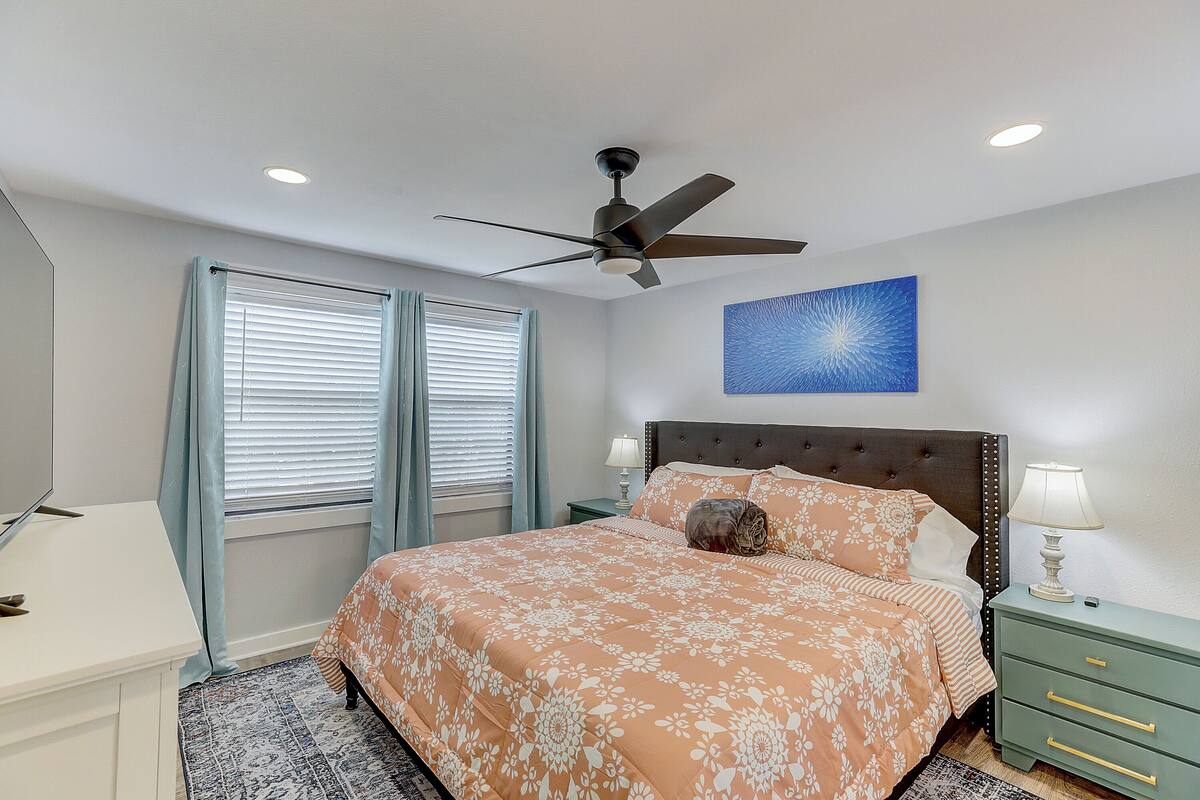 A bedroom with a bed, a ceiling fan, a nightstand with a lamp, and a wall-mounted artwork.