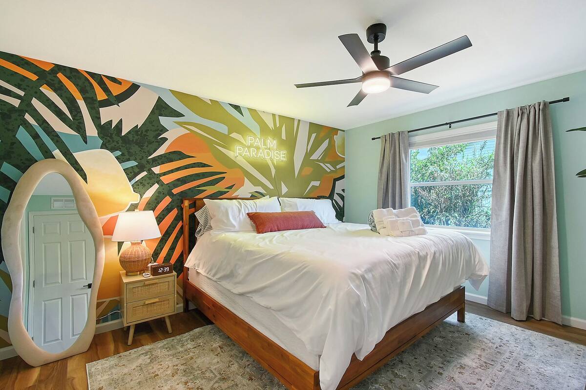 A bedroom with a bed, a nightstand, a lamp, a ceiling fan, a window with curtains, and a wall mural.