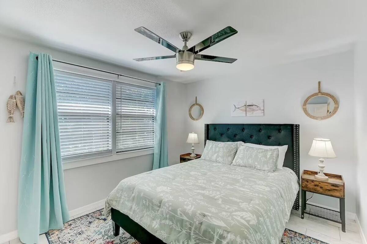 A bedroom with a bed, a ceiling fan, a window with blinds, and a nightstand with a lamp.