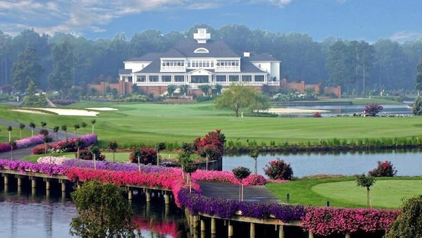 Een groot gebouw met een symmetrische gevel, een golfbaan op de achtergrond en een waterpartij met bloemenpaden.