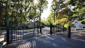 Een omheind toegangsweg met een metalen poort, omringd door bomen en een wit gebouw op de achtergrond.