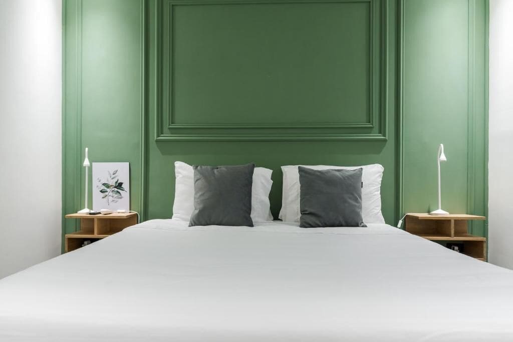 Una cama con ropa de cama blanca y dos almohadas, flanqueada por mesitas de noche con lámparas, frente a una pared verde con un panel empotrado.