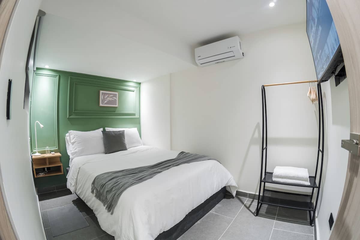 A bedroom with a bed, a green headboard, a wall-mounted air conditioner, a TV, and a towel rack.