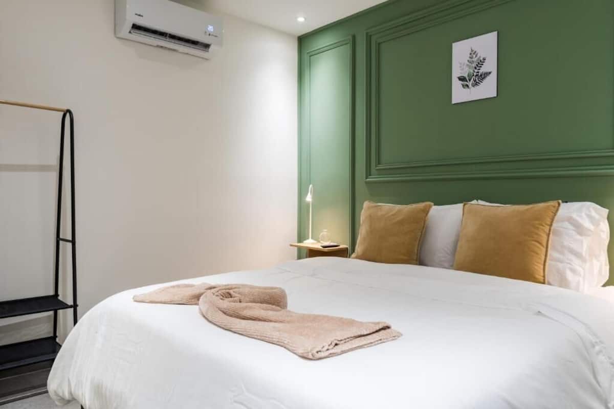 A neatly made bed with pillows, a bedside table with a lamp, and a wall-mounted air conditioner.