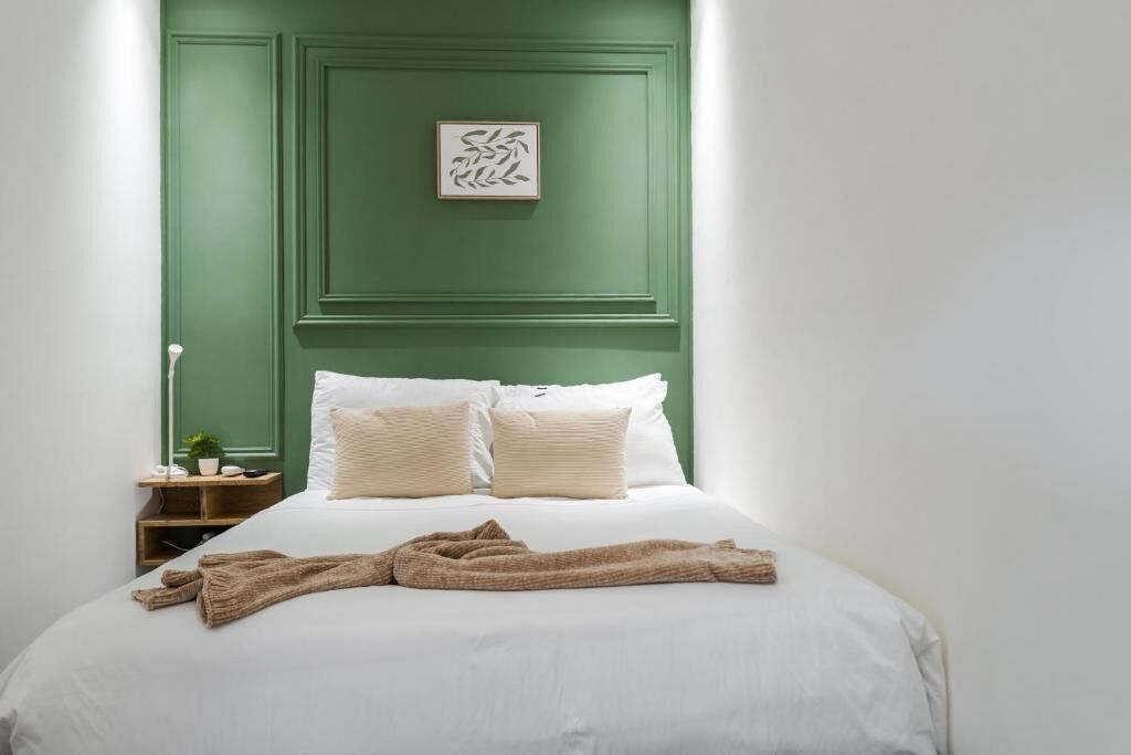 A neatly made bed with white linens and two pillows, a green accent wall, a framed artwork, and a bedside table with a lamp.