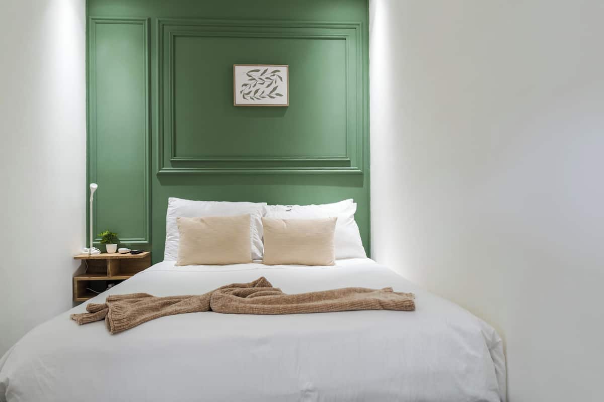 A neatly made bed with white linens and two pillows, a brown throw blanket, a bedside table with a lamp, and a framed artwork on the wall.