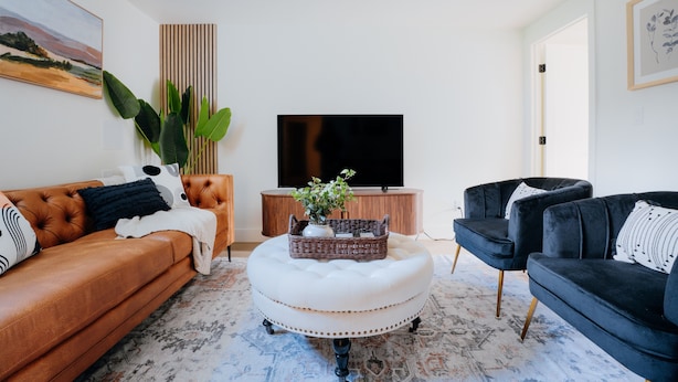 A living room with a brown leather sofa, a dark blue velvet sofa, a white ottoman, a flat-screen TV, and a large plant. - Sleeps 12 · 3-Kitchen · Walk to Link Light Rail (Seattle)
