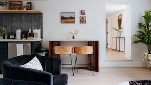 A modern living room with a black sofa, a coffee machine, a small dining table with two stools, and a large plant. - Sleeps 12 · 3-Kitchen · Walk to Link Light Rail (Seattle)