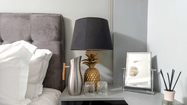 A neatly made bed with white linens, a grey upholstered headboard, a modern lamp with a black shade, a decorative pineapple vase, two clear glass vases, a framed mirror, and a small black bag on a white nightstand. - The Fox at Hurley - The Warren Suite (Maidenhead)