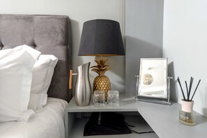 A neatly made bed with white linens, a grey upholstered headboard, a modern lamp with a black shade, a decorative pineapple vase, two clear glass vases, a framed mirror, and a small black bag on a white nightstand. - The Fox at Hurley - The Warren Suite (Maidenhead)