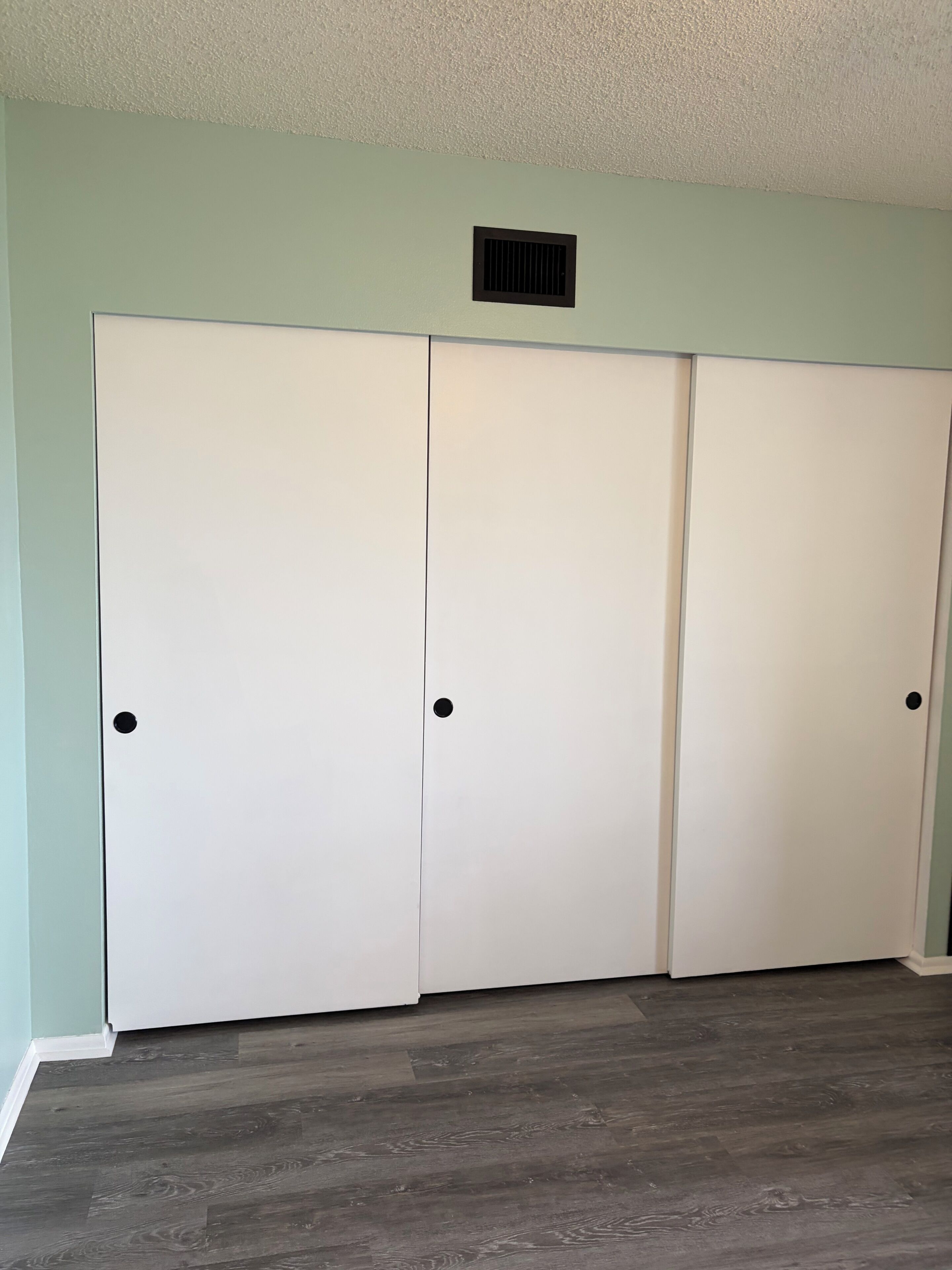 A room with a light green wall, a white closet with two doors, and a vent above the closet.