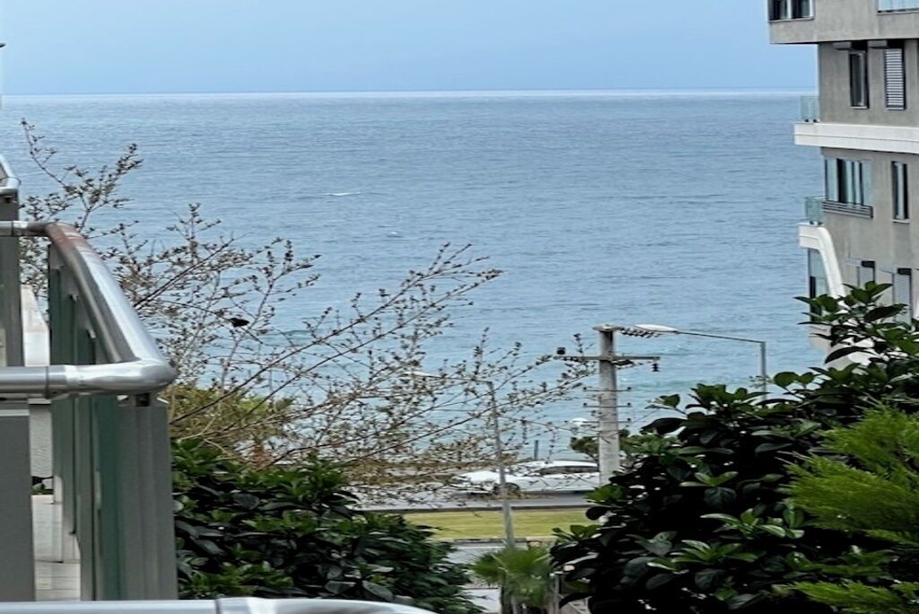 A coastal building with a balcony overlooking the sea, surrounded by greenery.