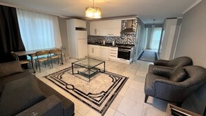 Een moderne woonkamer met een glazen salontafel, een bank, een eethoek met tafel en stoelen, een keuken met witte kastjes en een zwart aanrecht, en een gang die naar een andere kamer leidt.