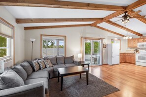 Un vaste espace de vie comprenant un canapé d’angle, une table basse, des poutres apparentes, un ventilateur de plafond et une cuisine dotée de meubles et d’appareils électroménagers en bois.