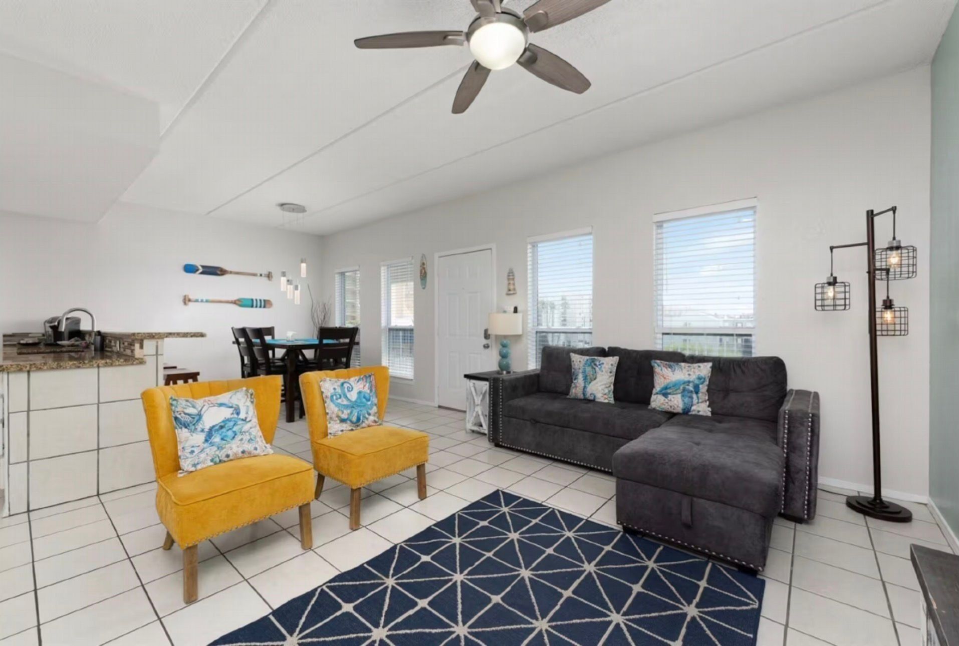 A modern living room with a gray sofa, two yellow armchairs, a dining area, and a ceiling fan.