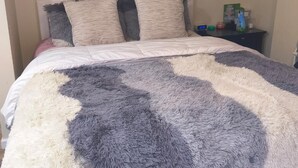 Uma cama com um cobertor fofo, uma mesa de cabeceira com um relógio e uma pequena mesa com artigos de higiene pessoal.