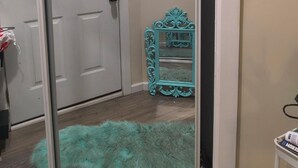 Um corredor com porta branca, espelho decorativo azul-turquesa e tapete felpudo no chão.