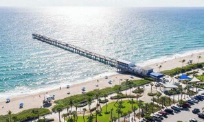 Um cais que se estende para o oceano, com uma praia e área de estacionamento ao primeiro plano.