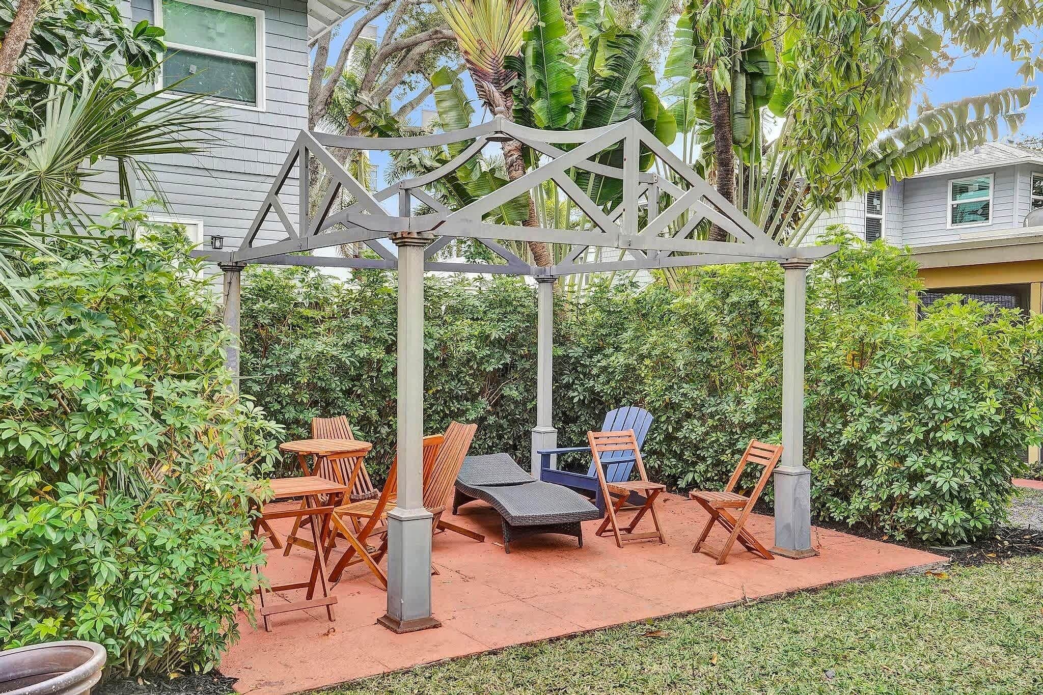 Un área de estar al aire libre techada con sillones y una mesa, rodeada de follaje y una casa al fondo.