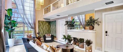 A spacious living area with a sofa, a round coffee table, and a fireplace. There are large windows with sheer curtains, a balcony with decorative railings, and various potted plants.