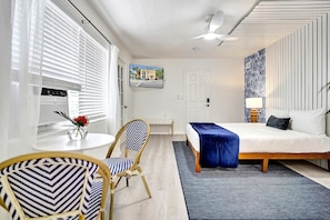 A modern hotel room with a bed, a desk, a chair, a radiator, and a wall-mounted TV. - Renovated Studio | Walk to Downtown Lake Worth (Lake Worth Beach)