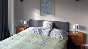 Ett sovrum med en säng, två sängbord, ett litet bord, en inramad tavla och ett fönster med gardiner.