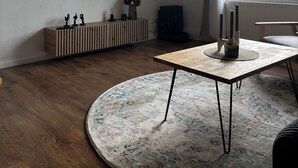 Ett modernt vardagsrum med en platt-TV som är monterad på väggen, ett träbord, en rund matta och ett fönster med utsikt över omgivningen.