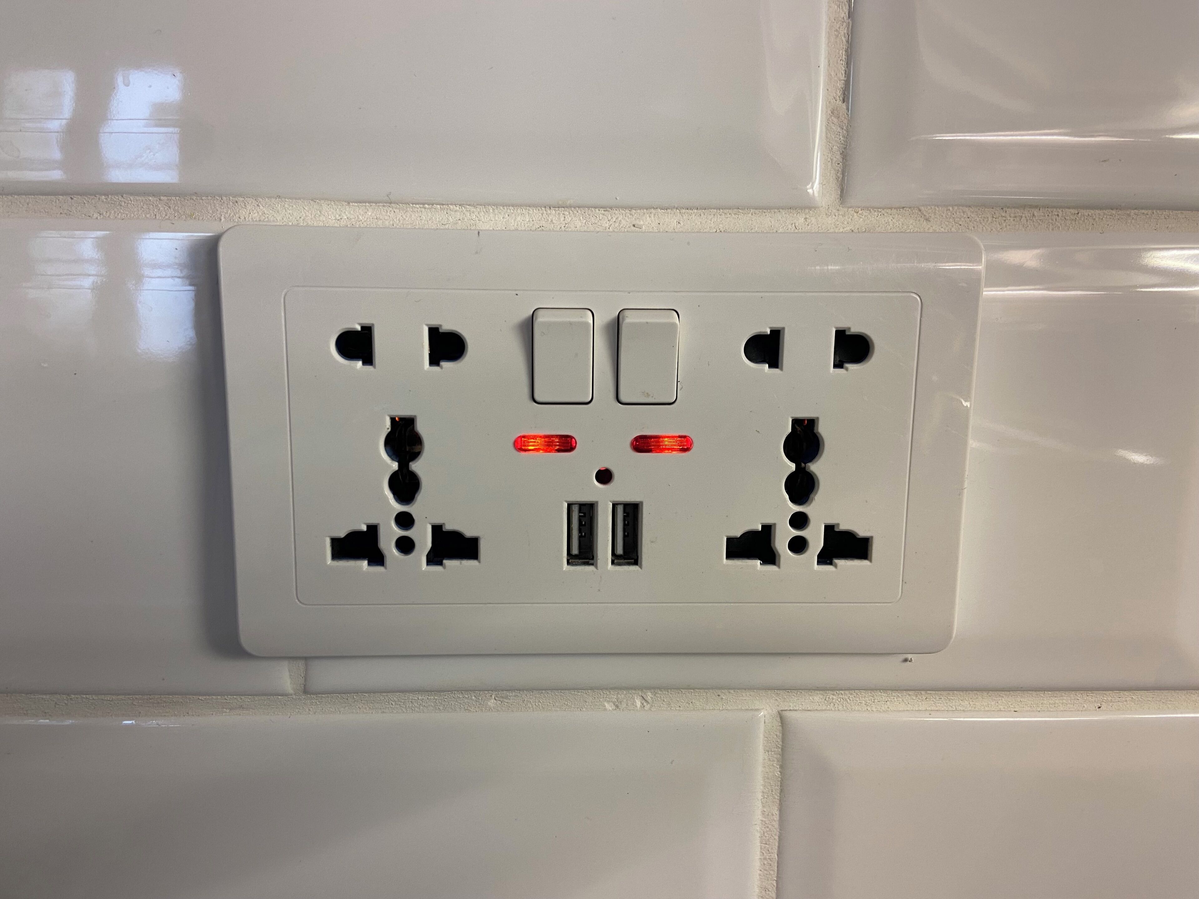 A white electrical outlet with two switches and two red indicator lights on a tiled wall.
