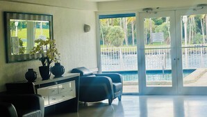Une pièce comprenant un bureau, deux chaises, une grande fenêtre donnant sur une piscine et sur de la verdure, ainsi qu’un miroir au mur.