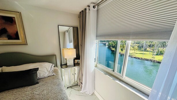 Une chambre à coucher comprenant un lit, une table de chevet, un miroir et une fenêtre donnant sur un plan d’eau et de la verdure.