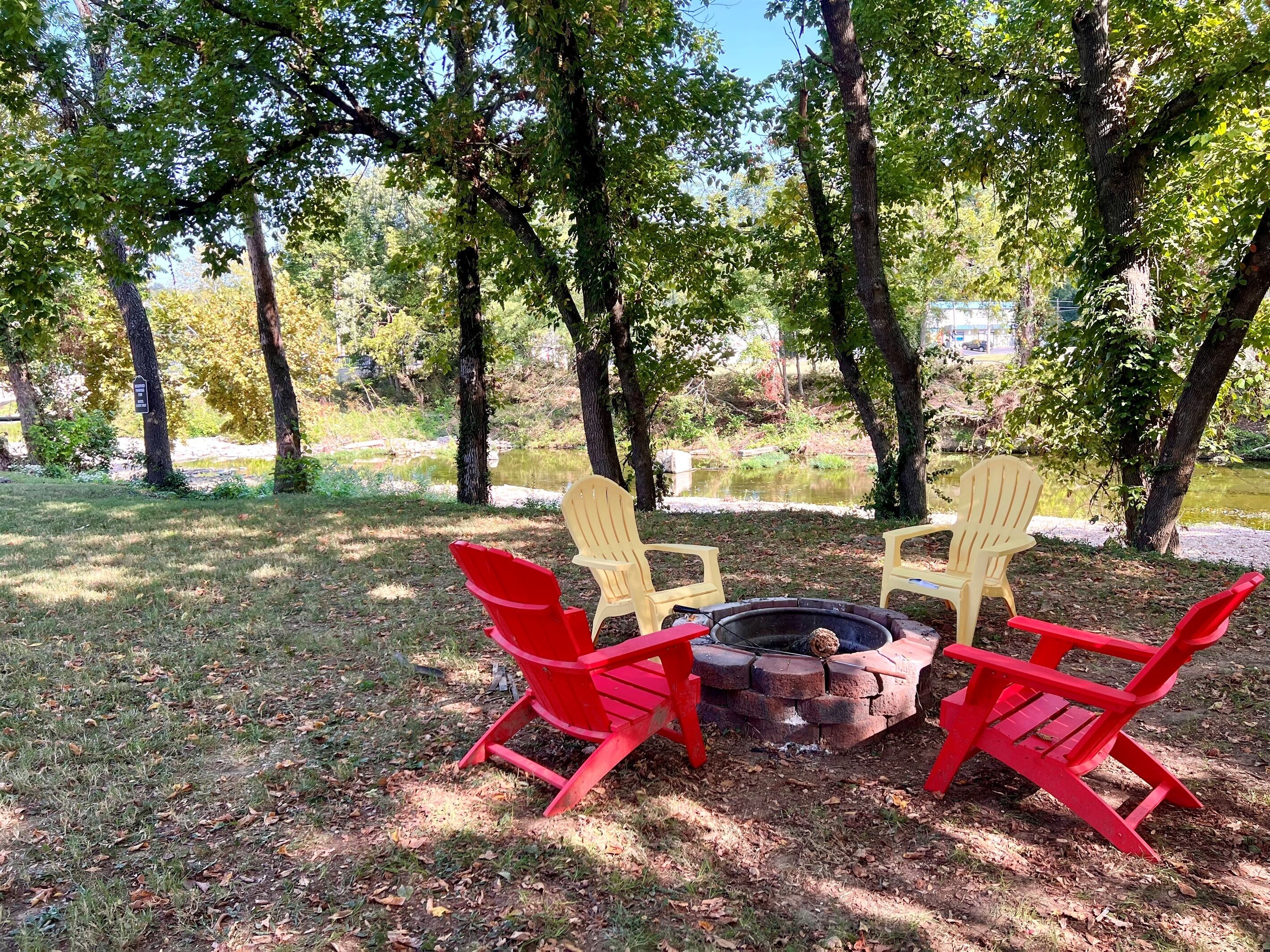 A fire pit surrounded by four Adirondack chairs, with trees and a grassy area in the background.