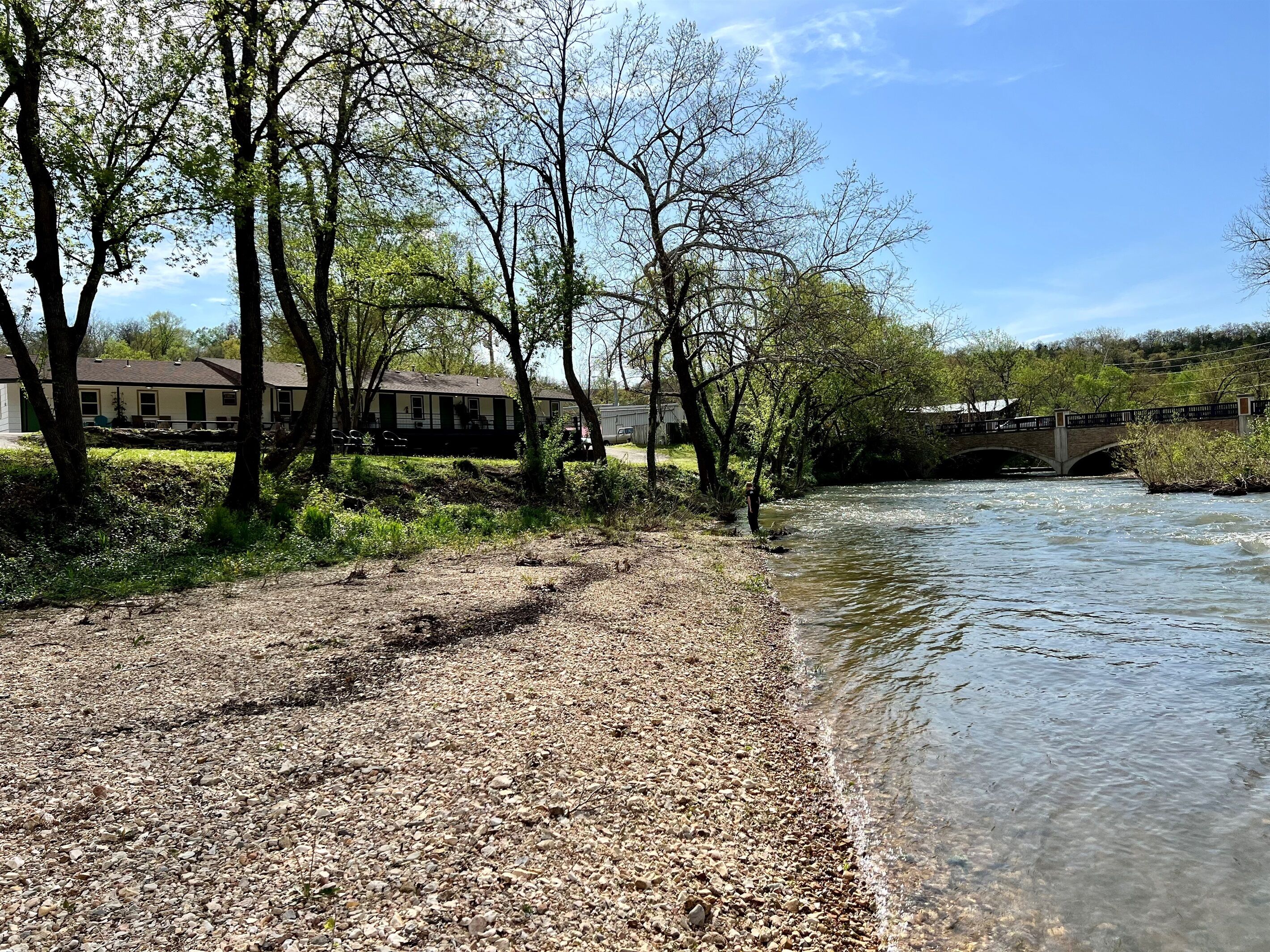 A riverbank with a bridge, trees, and a building in the background.