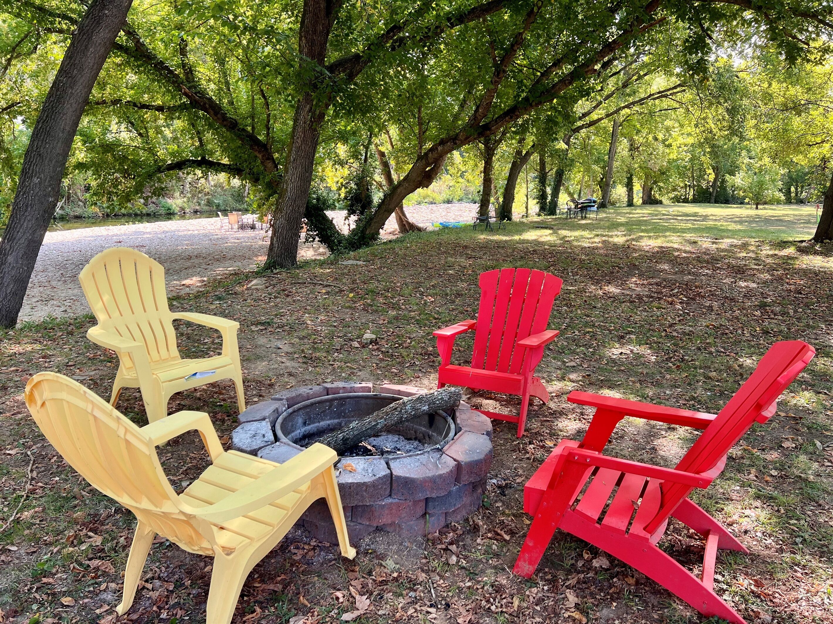 A fire pit with four Adirondack chairs, surrounded by trees in a park setting.