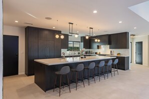 A modern kitchen with dark cabinetry, a central island with bar stools, recessed lighting, and a large window. - Casa Dona by Oraklus (Beverly Hills)