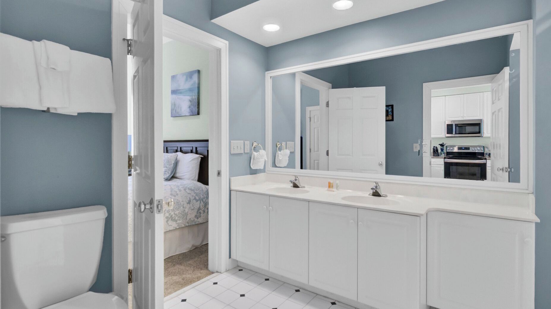 A bathroom with a double sink, a large mirror, and a door leading to a bedroom.