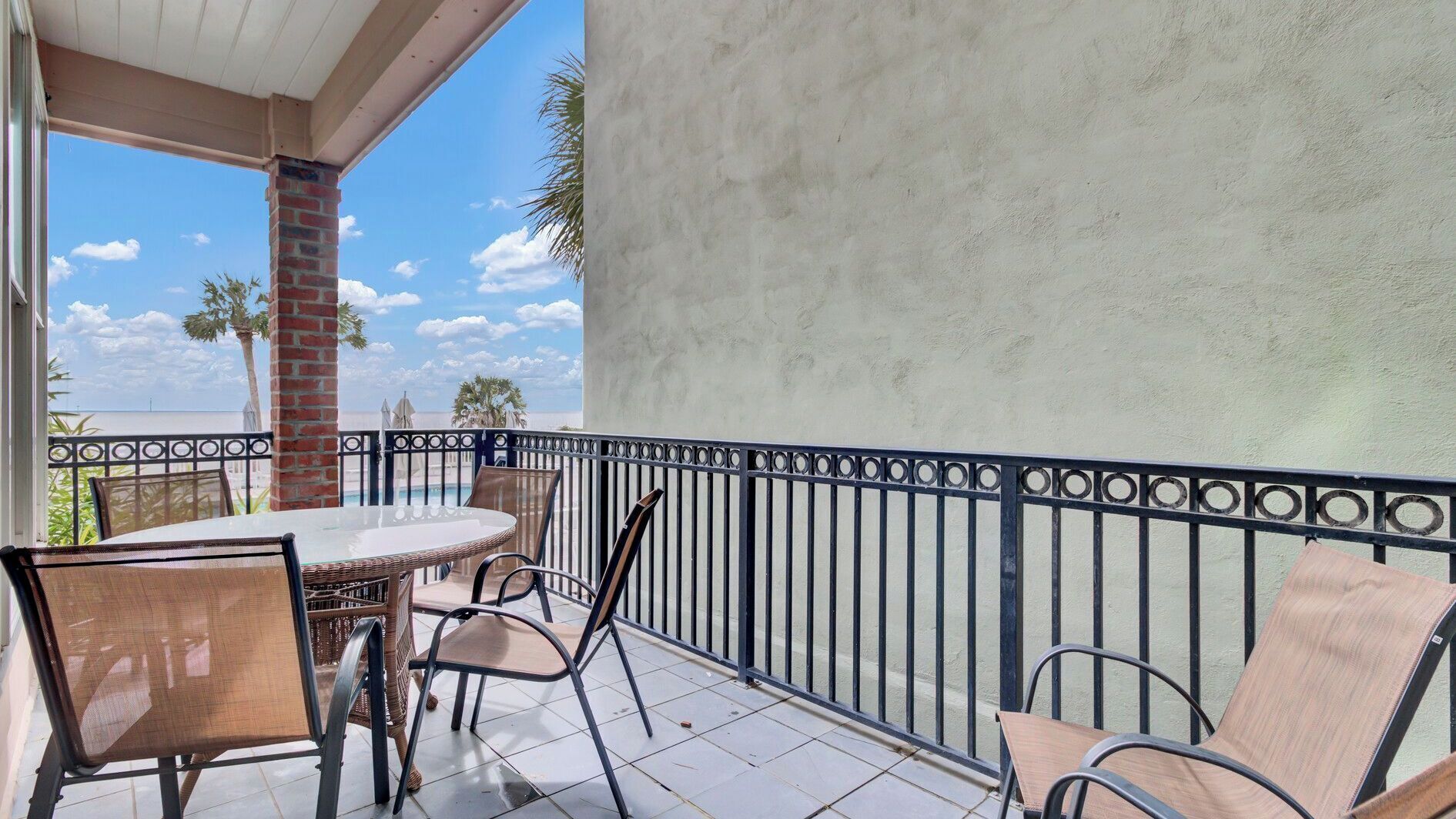 A balcony with a table and chairs, a brick pillar, and a view of the beach and palm trees.