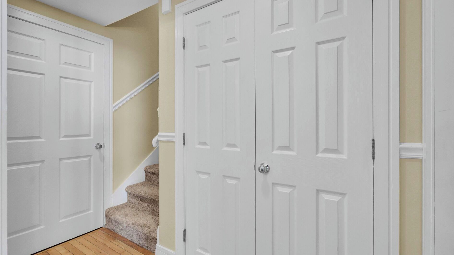 A hallway with white doors, a carpeted staircase, and a beige wall.