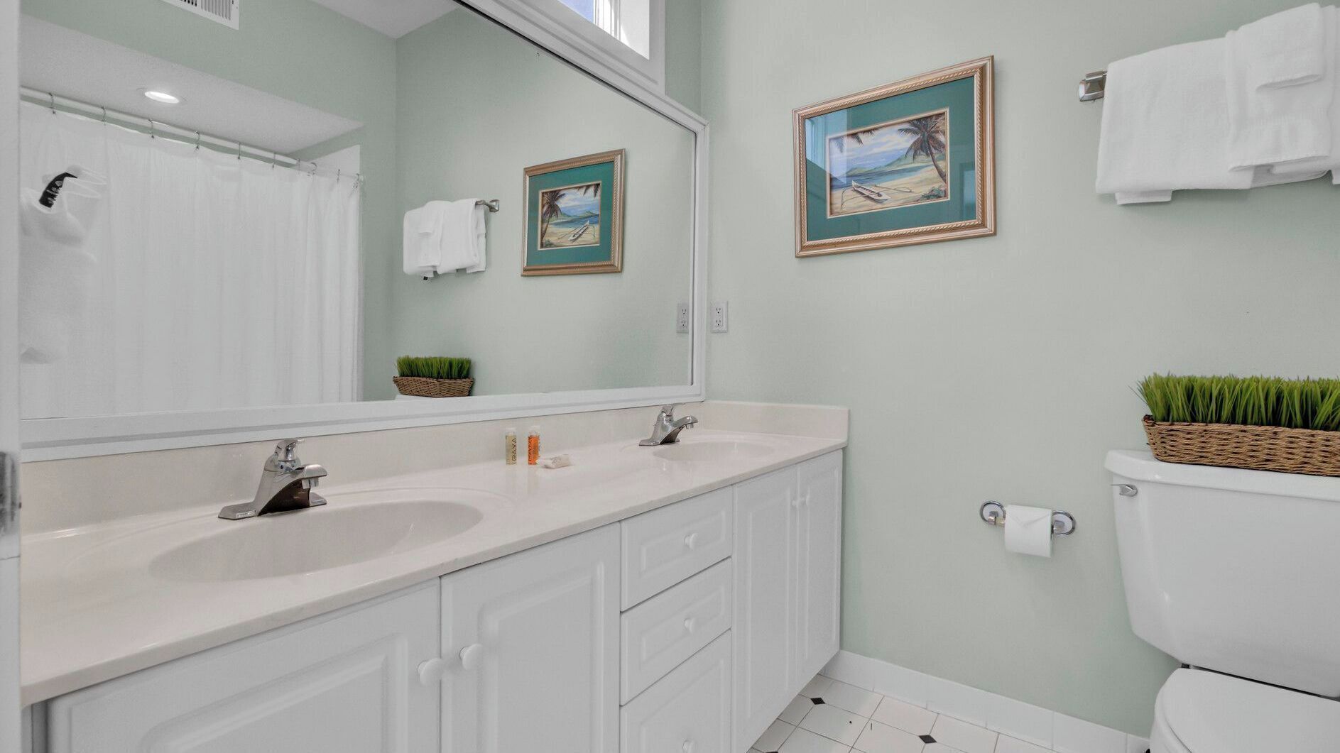 A bathroom with a large mirror, two sinks, a toilet, and framed pictures on the wall.