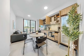 A modern kitchen with built-in appliances, a dining area with a round table and chairs, and a view of a cityscape through large windows. - Sleek 1-BR & Den in Miami Central - Rooftop Pool (Miami)
